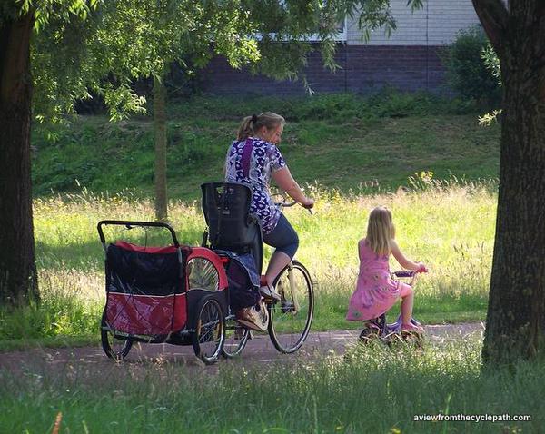 Mother cycling with daughter and with child-carry trailer behind